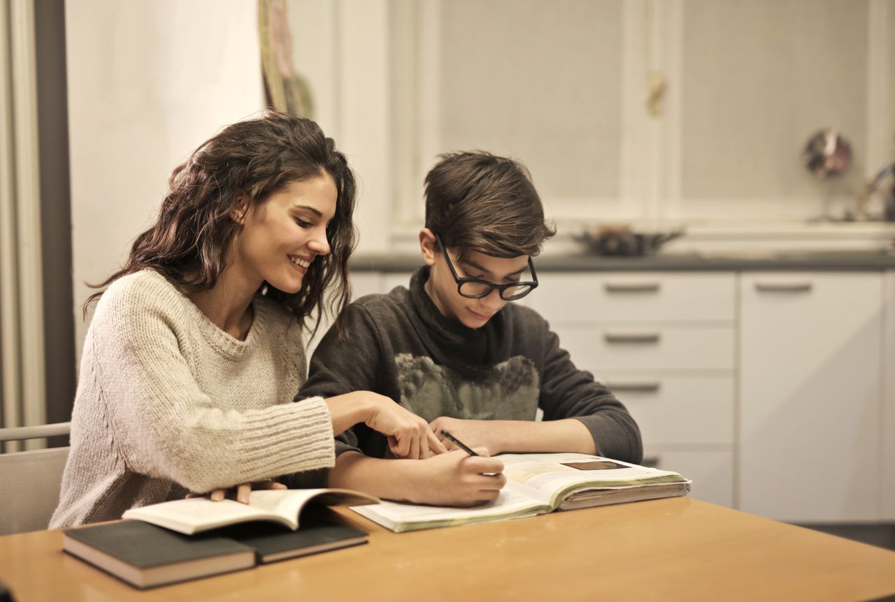 Cheerful young woman helping younger brother with homework while sitting together at table with books at home