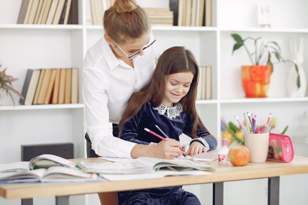 Young woman in casual clothes helping diligent girl to write in notebook while sitting at table with textbooks and doing homework in classroom at school