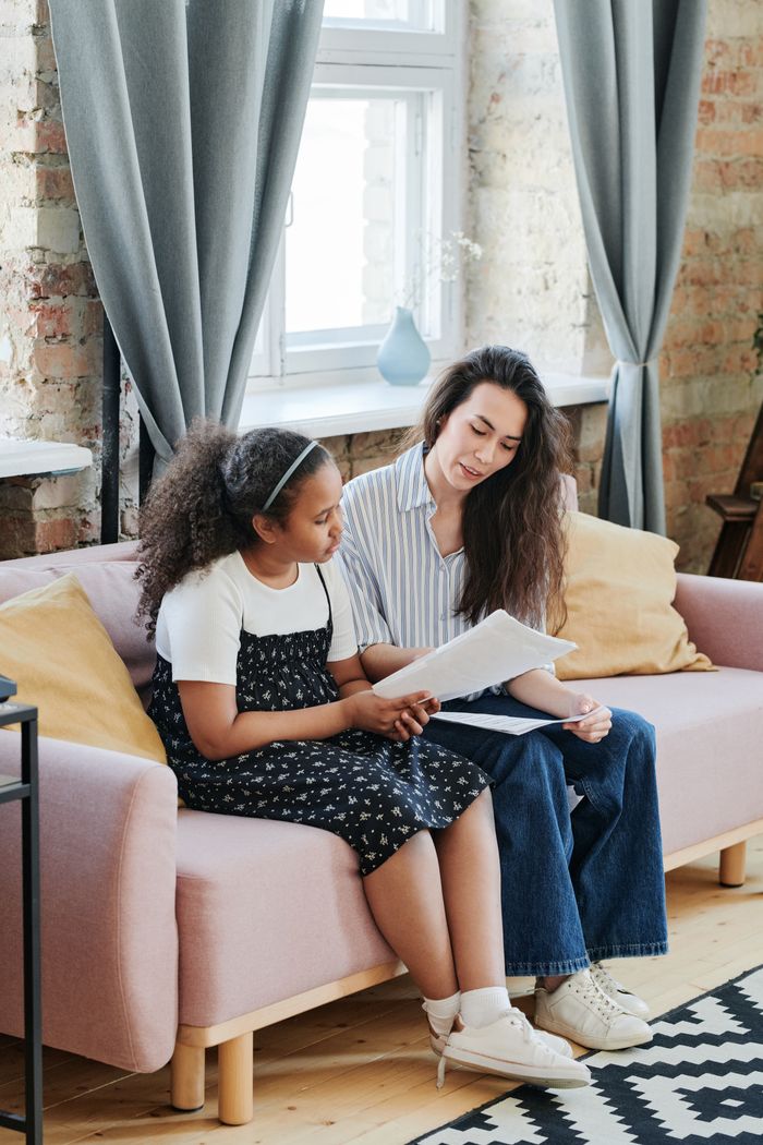 A woman and young girl sitting on a couch reading documents in a cozy living room.