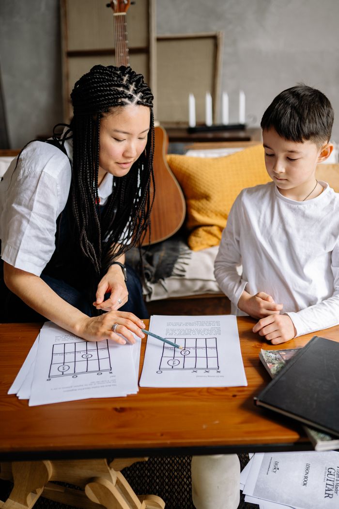 An adult teaching a child to read music notes on a sheet indoors.